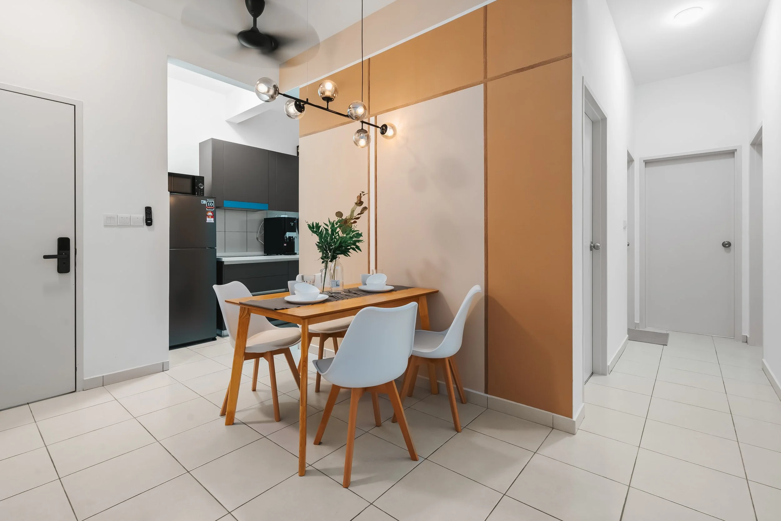 Modern dining area with a wooden table set for four and white chairs, adjacent to a kitchen with black cabinets and a stainless steel refrigerator, decorated with a green plant, pendant lights, and white tiled flooring.