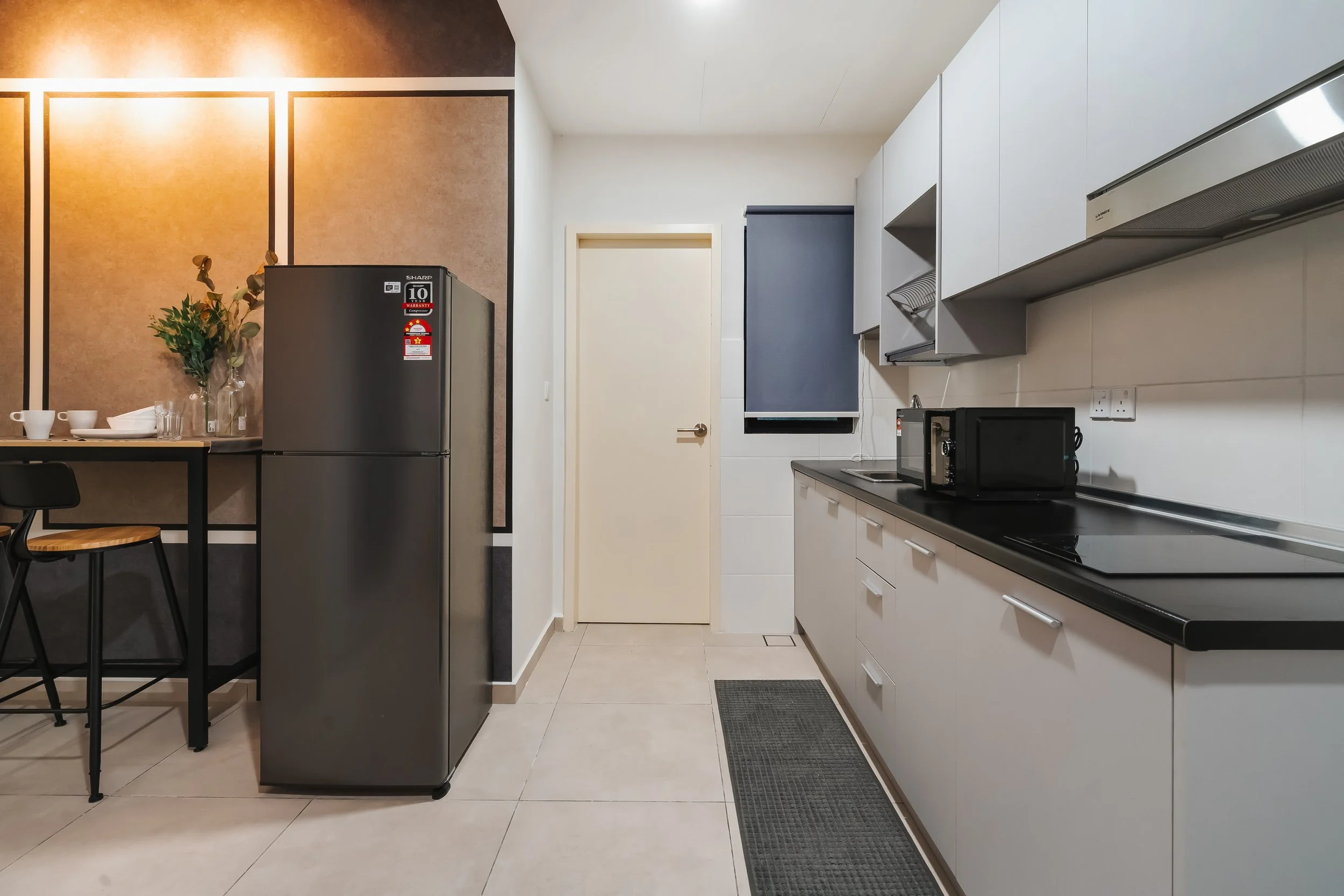 Modern kitchen with white cabinets, black countertop, microwave, and range hood. A door and window with a blue blind are visible, alongside a small table with chairs, a fridge, and decorative items.