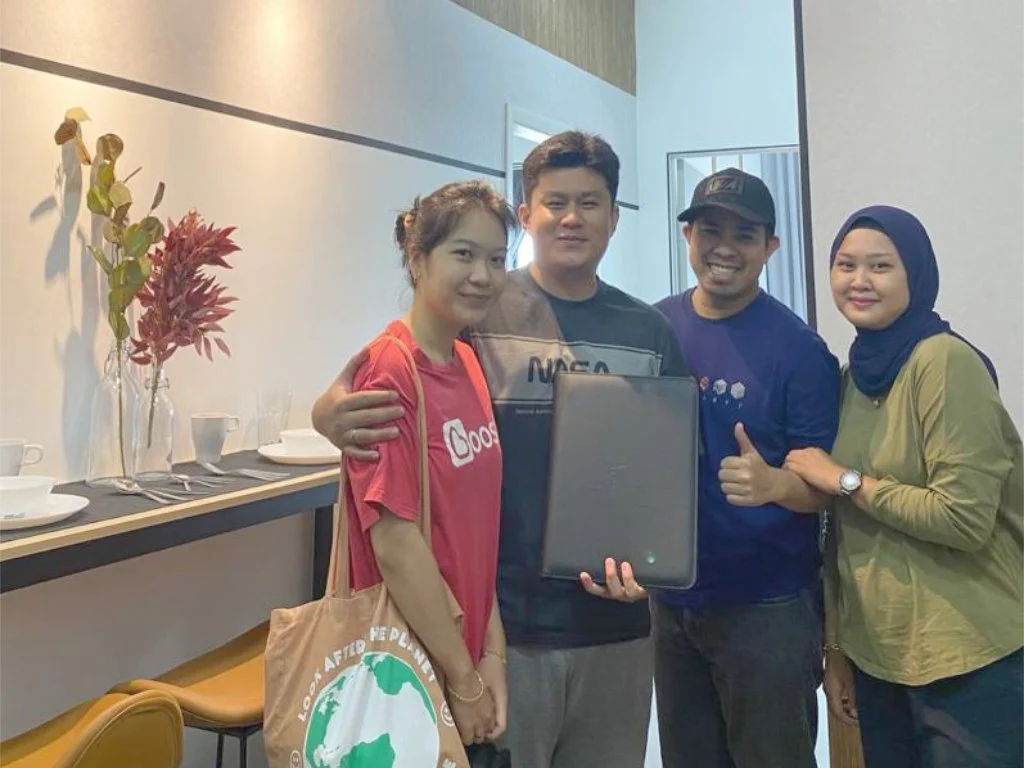 Group of four people smiling, with one holding a laptop in a room with minimal decor and a table with plates and cups