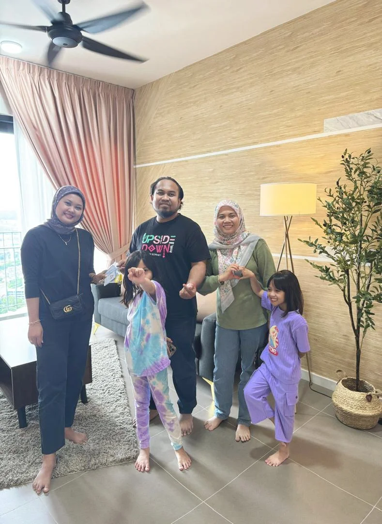 Group of five people, three women and two children, standing in a living room near a window with pink curtains, smiling at the camera. The women are wearing modest clothing and hijabs. The children are dressed in colorful pajamas, and all are barefoo