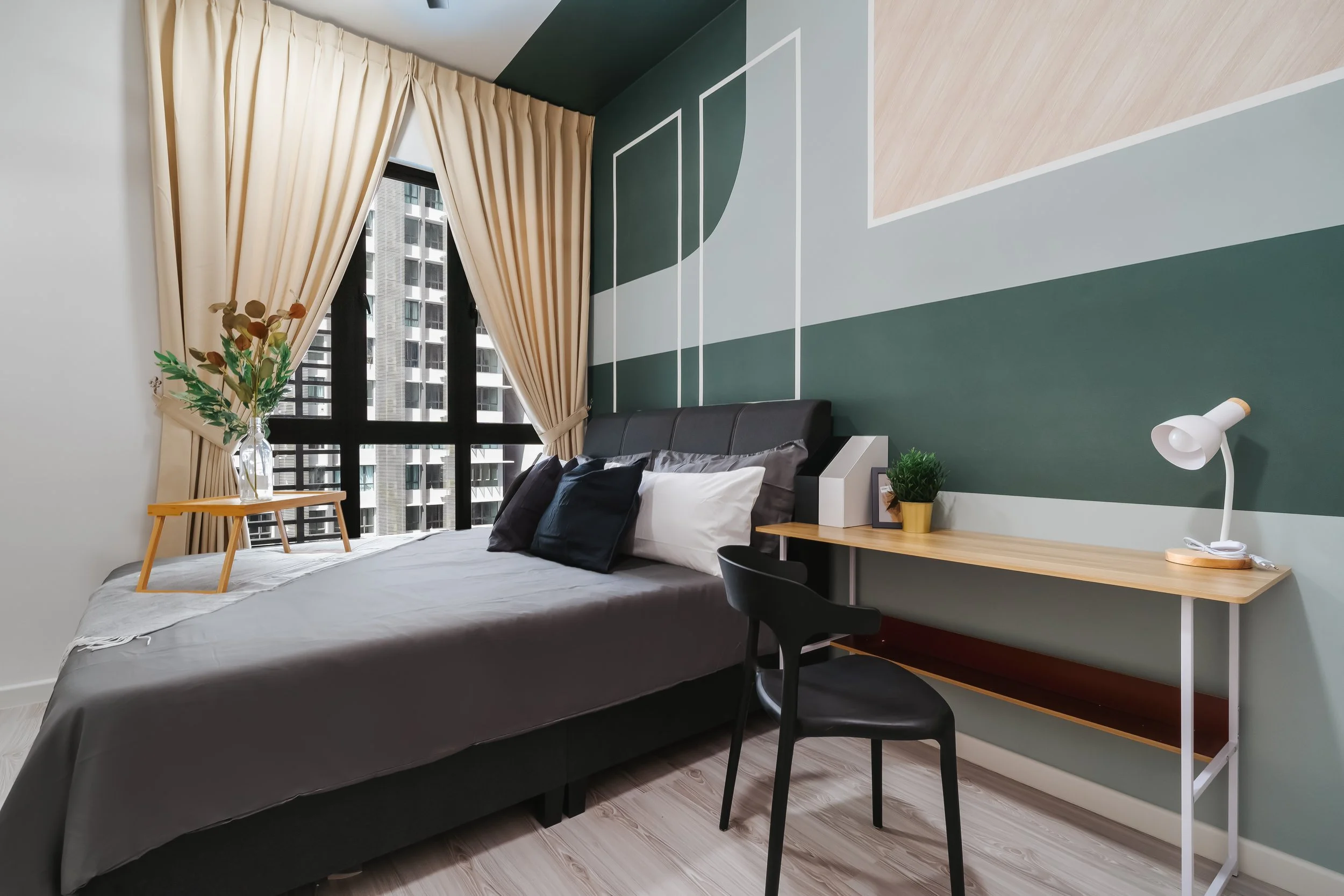 A modern bedroom with a large bed, black headboard, gray bedding, and pillows. A wooden side table with a vase holding greenery is next to the window. The window has beige curtains and a city view with tall buildings. The wall behind the bed has geom