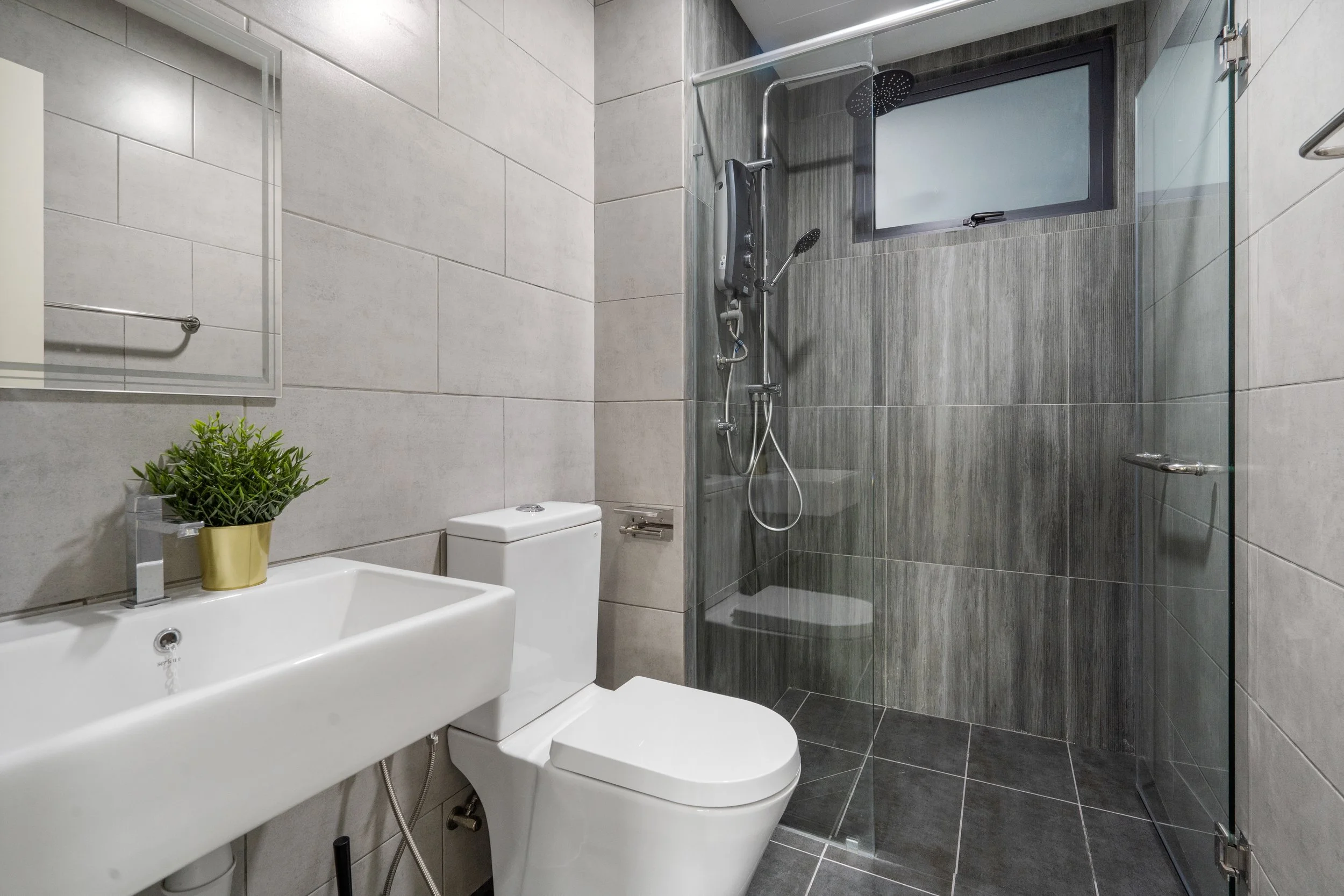 Modern bathroom featuring a white toilet, a rectangular sink with a green potted plant, a large mirror, and a walk-in shower with glass walls and a black showerhead, with gray tiled walls and floors.