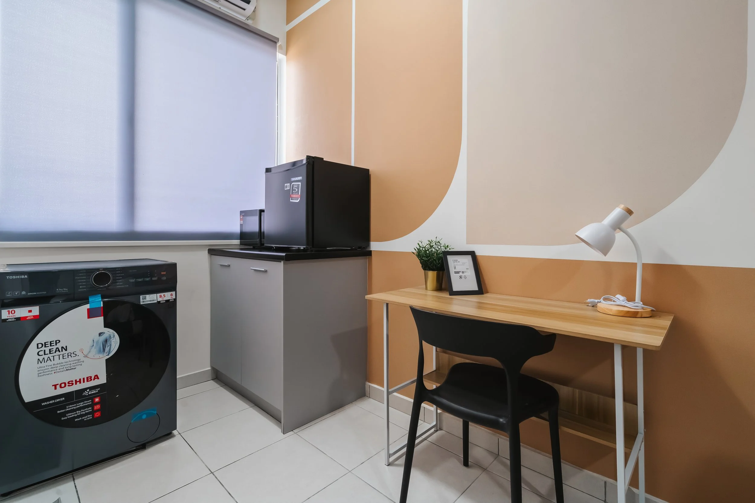 Laundry room with white tiled floor, a washing machine with a detergent sticker, a black mini fridge on top of a grey cabinet, a wooden table with a black chair, a white desk lamp, a small green plant in a gold pot, a picture frame, and a window with