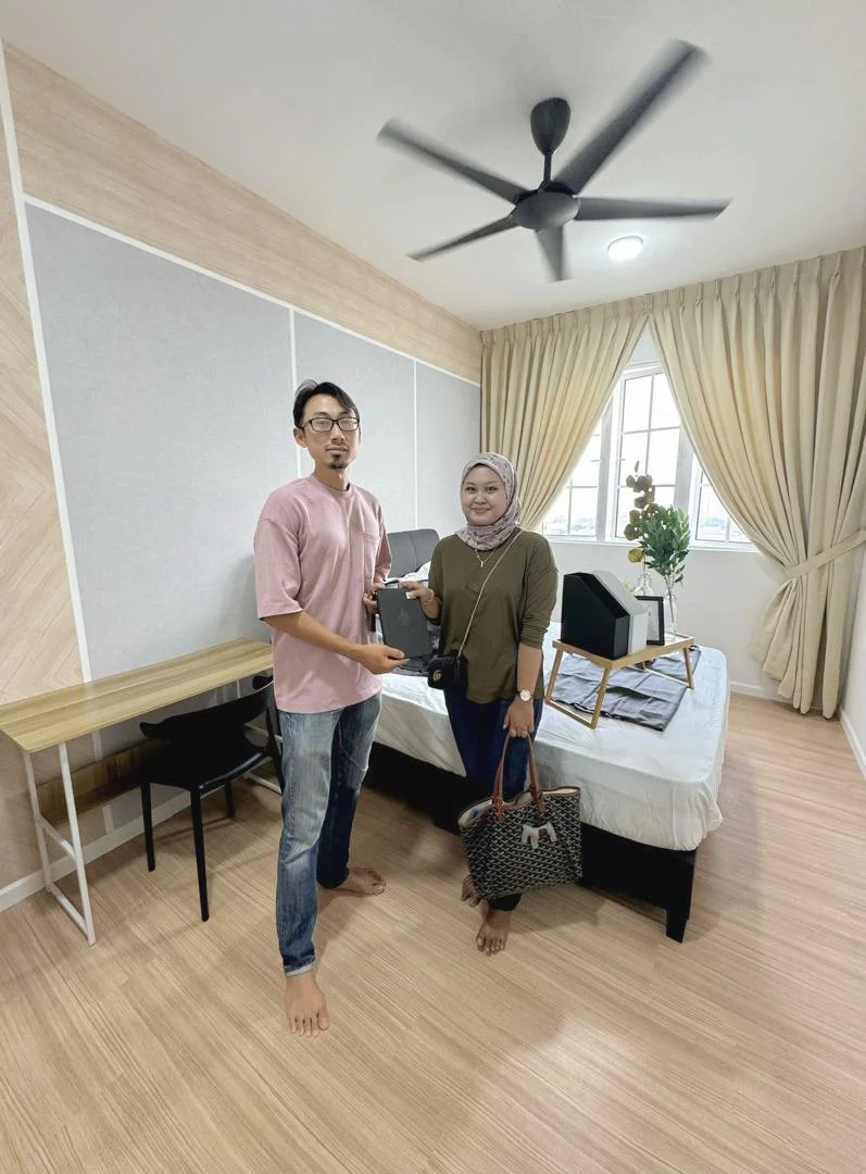 A man and a woman standing in a bedroom, holding a black box together. The woman is wearing a headscarf and carrying a patterned handbag. The man is dressed casually in a pink shirt and jeans. The room has a bed, a window with beige curtains, a woode