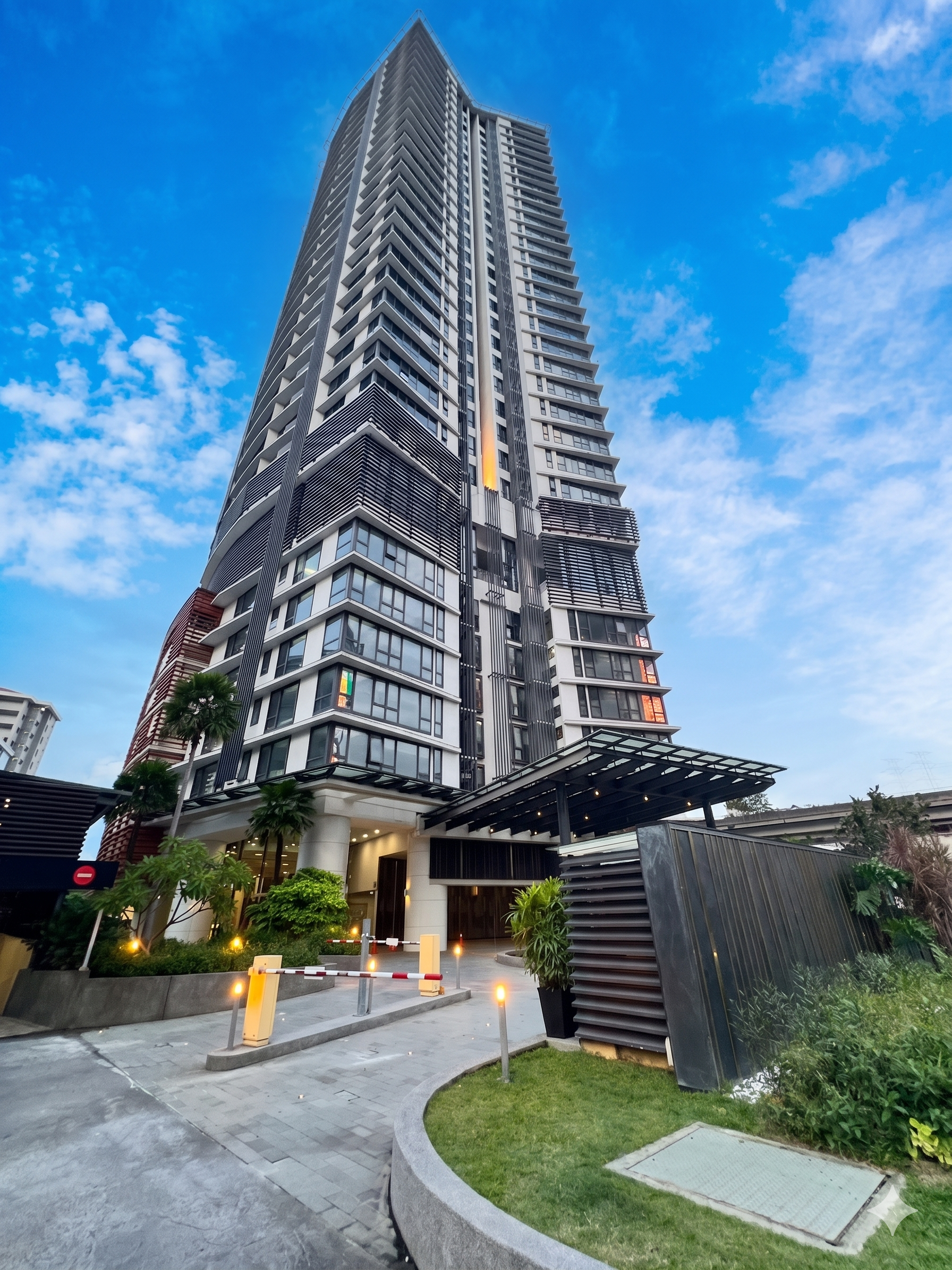 A tall modern high-rise building with a unique curved design, set against a bright blue sky with scattered clouds, with some greenery and a driveway at the base.