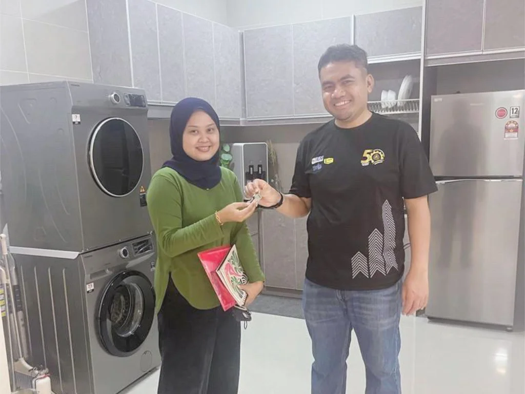 A woman wearing a green top and a dark headscarf receiving a key or small object from a smiling man in a black T-shirt, in a laundry or utility room with washing machines and a refrigerator.