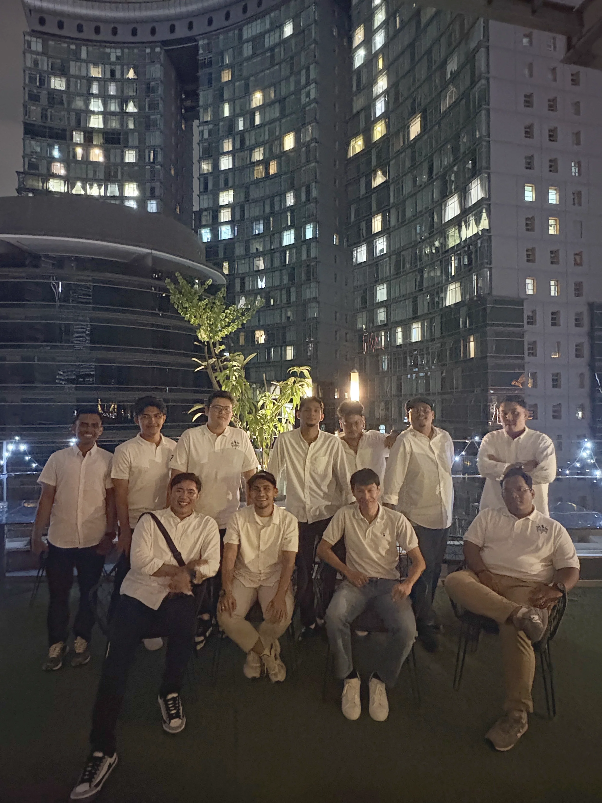 A group of people dressed in white shirts and beige or black pants, standing in an outdoor area at night with illuminated high-rise buildings in the background.