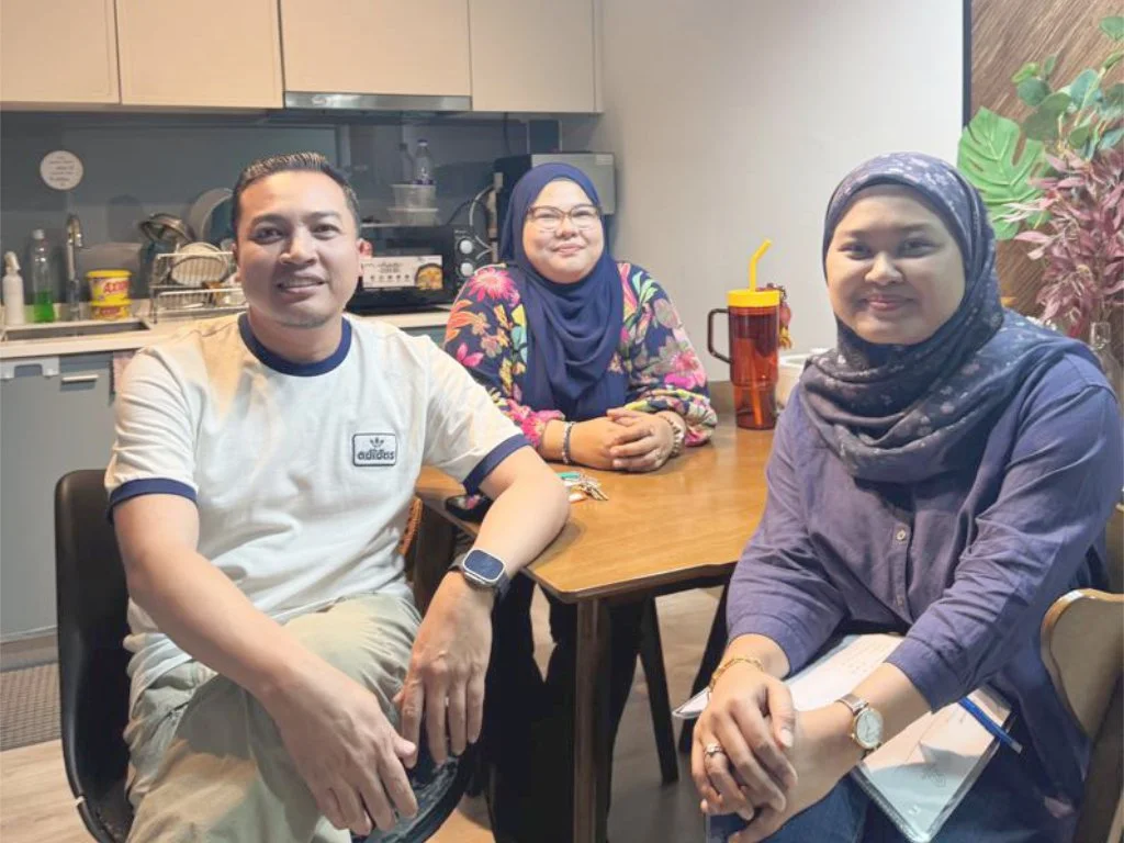 Three people sitting at a wooden table in a kitchen, smiling at the camera. Two women wearing headscarves sit on the right and center, while a man in a white t-shirt and khaki pants sits on the left. There are dishes and kitchen items in the backgrou