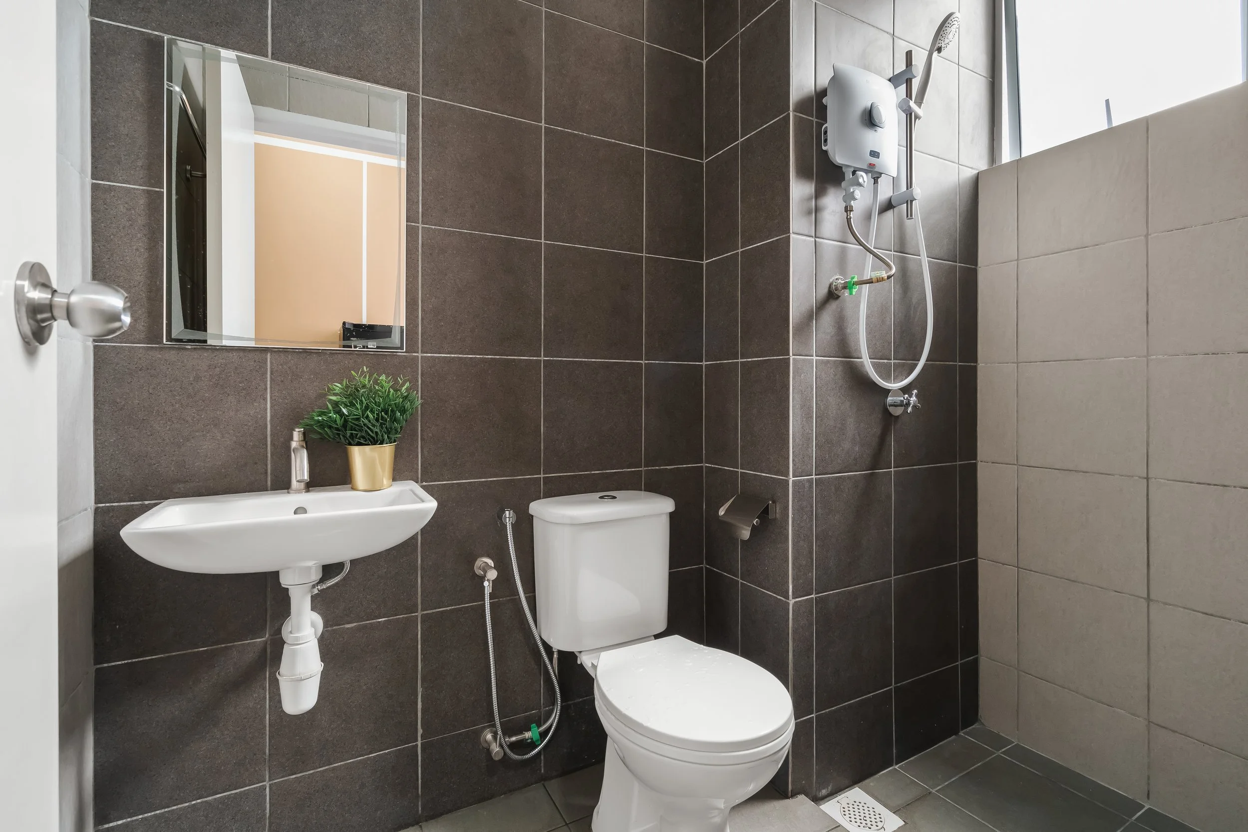 A modern bathroom with dark gray tiled walls and floor. It features a small white wall-mounted sink, a white toilet, a mirror above the sink, and a potted plant on the sink. There is an electric water heater and a handheld showerhead on the wall, and