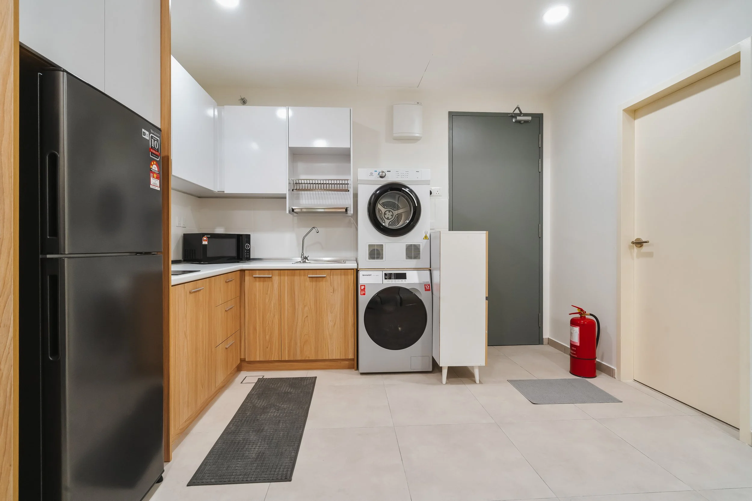 A laundry room with white walls and tile flooring, equipped with a stainless steel refrigerator, microwave, white cabinets, a sink, a washing machine, a dryer, and a fire extinguisher.