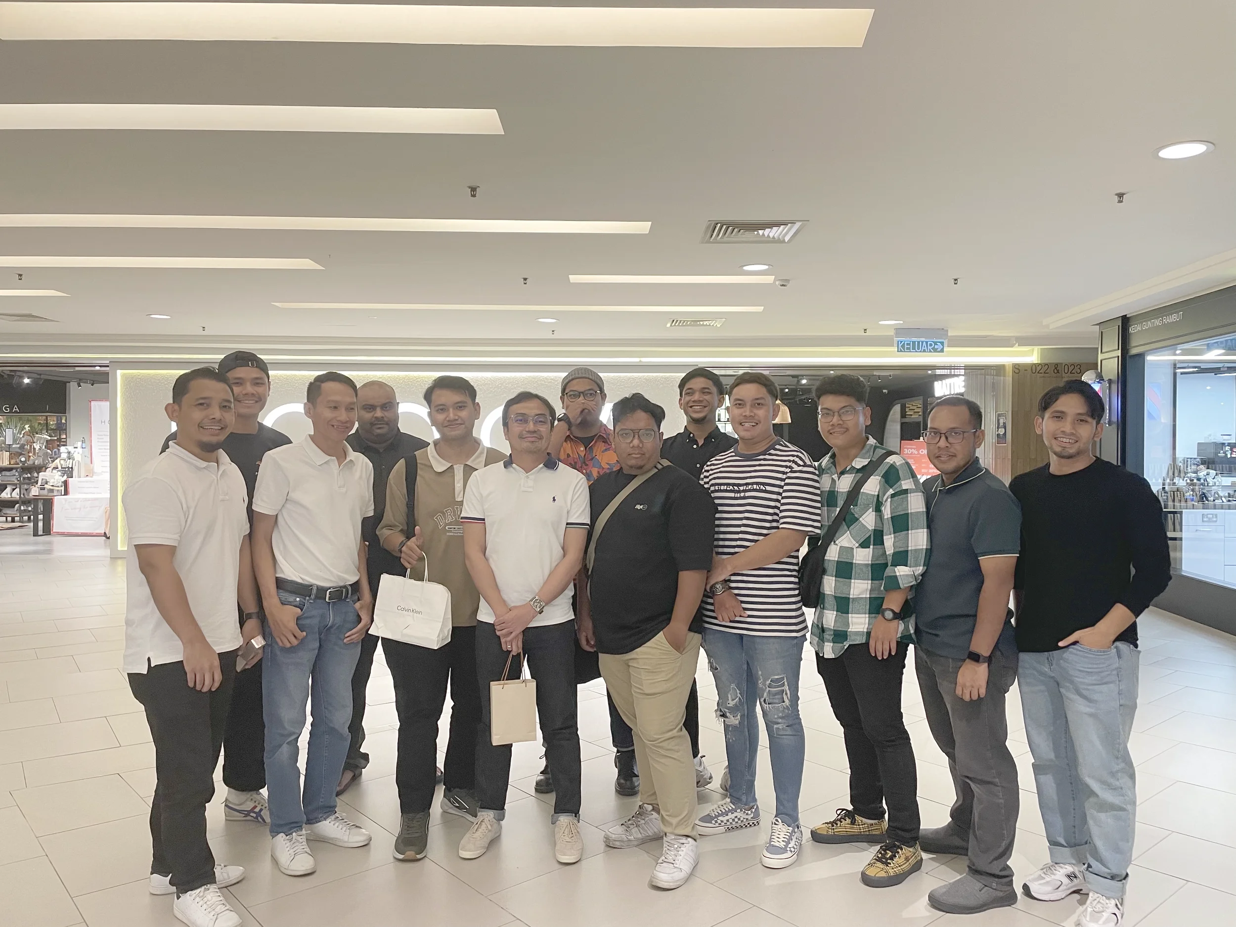 Group of young men and women standing together inside a shopping mall for a photo. They are smiling and casually dressed, with some wearing glasses and carrying shopping bags.