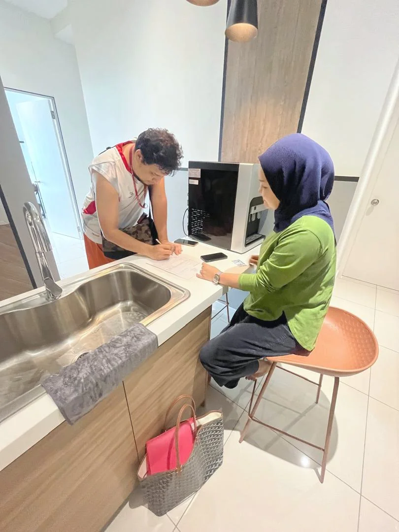 Two women are in a kitchen, one is signing papers at a counter while the other is seated on a chair, looking at her phone. There is a sink, a towel, and a shopping bag nearby. A desktop computer and some electronics are on the counter.