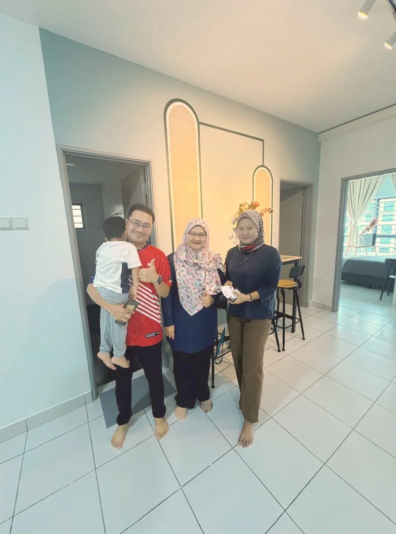 Four people standing indoors, two women and two men, with a young child. The man holding the child is wearing glasses and a red shirt. The women are wearing hijabs, and one woman is holding money. The background shows a modern, well-lit room with a l