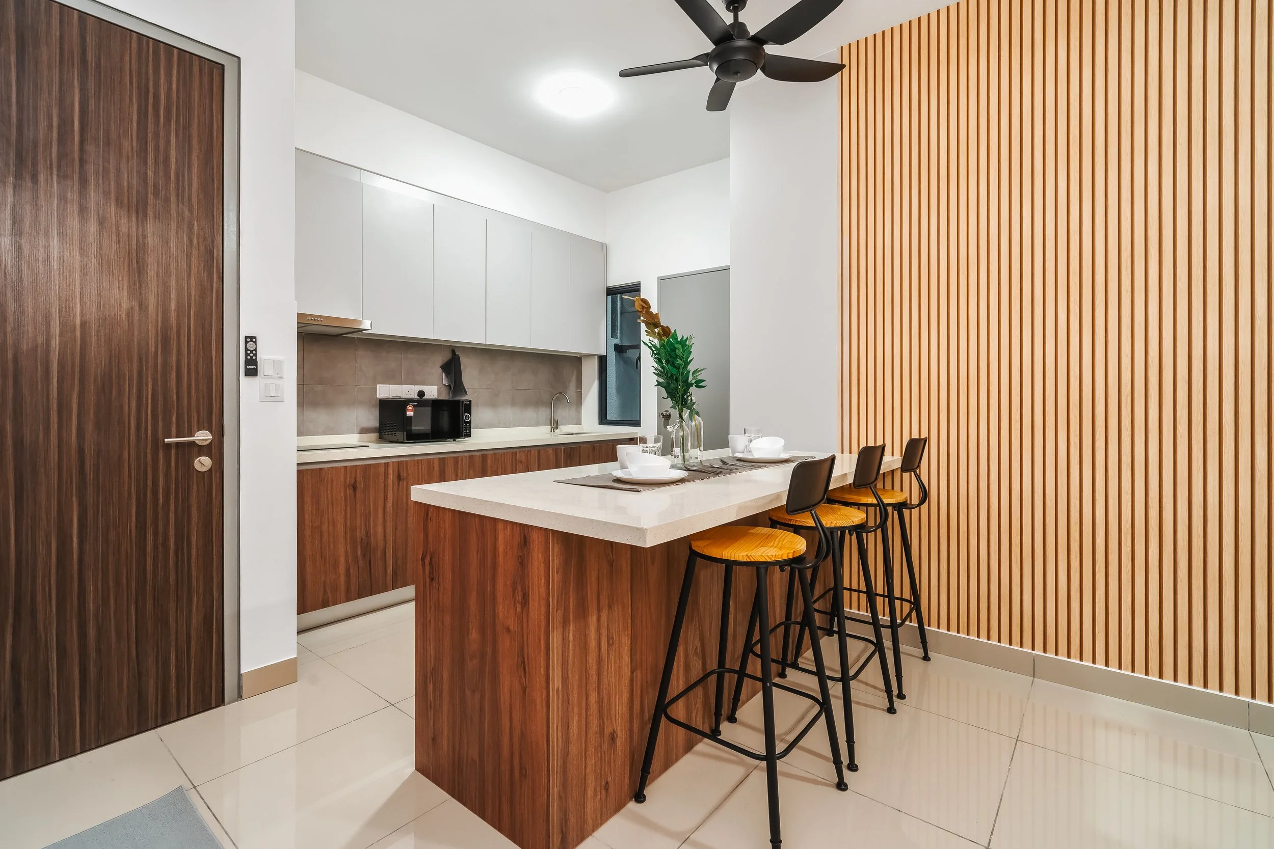 Modern kitchen with white upper cabinets, wooden lower cabinets, a private window, a microwave, and a dining area with a white marble countertop island, black stools with wooden seats, and a decorative green plant on the countertop.