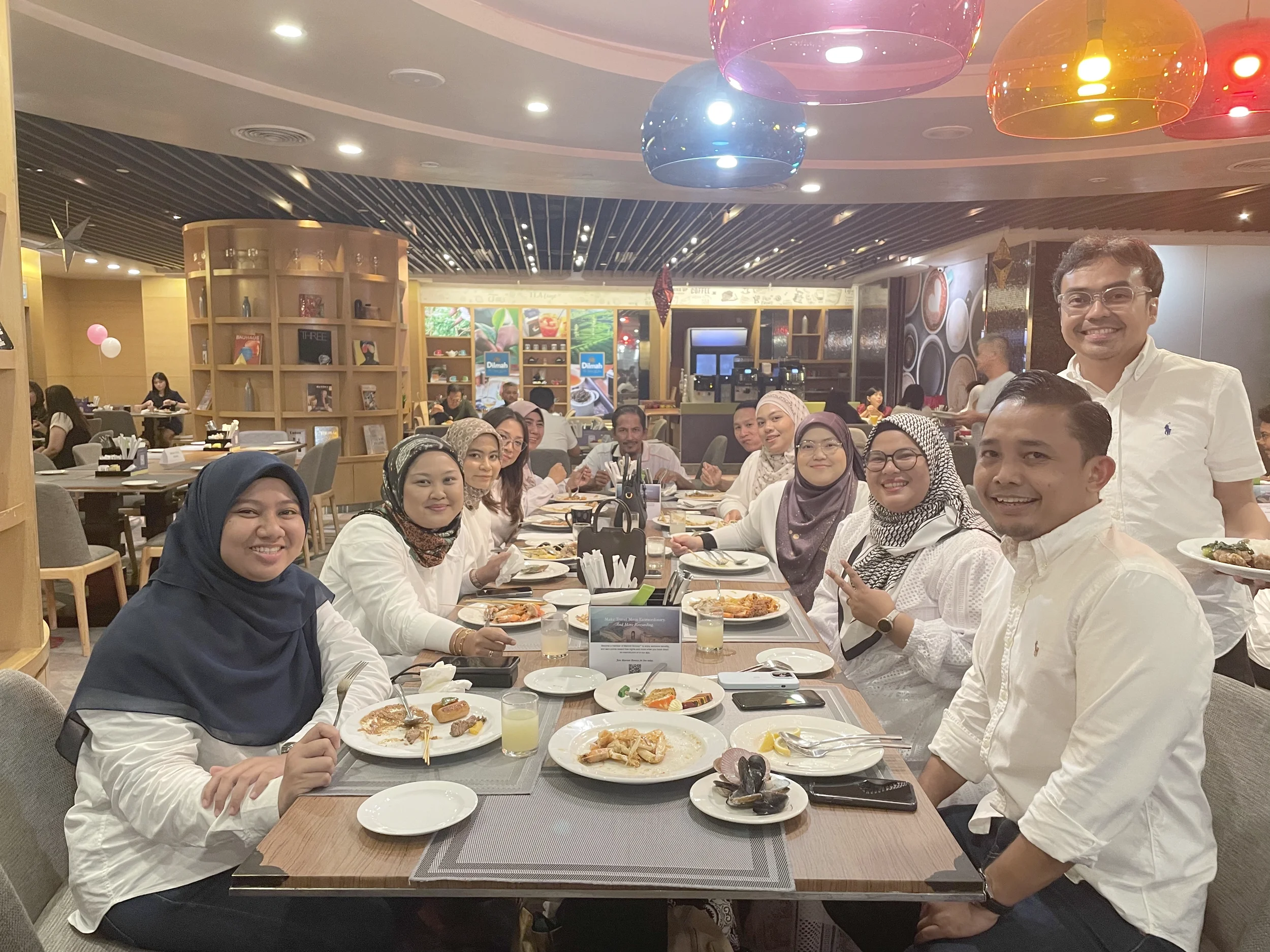 A group of people sitting around a restaurant table with plates of food, smiling for the camera, with a colorful ceiling and other diners in the background.