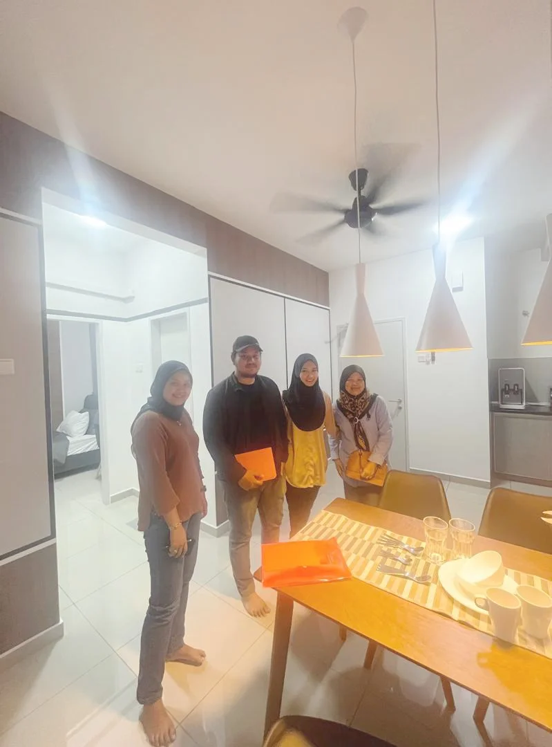 Four people standing inside a modern kitchen near a dining table, with pendant lights hanging from the ceiling.