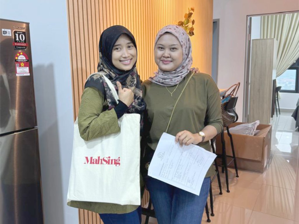 Two women wearing hijabs standing inside a room, smiling at the camera. One woman holds a shopping bag and the other holds a document. The background includes a wooden wall, a window, and some chairs and tables.