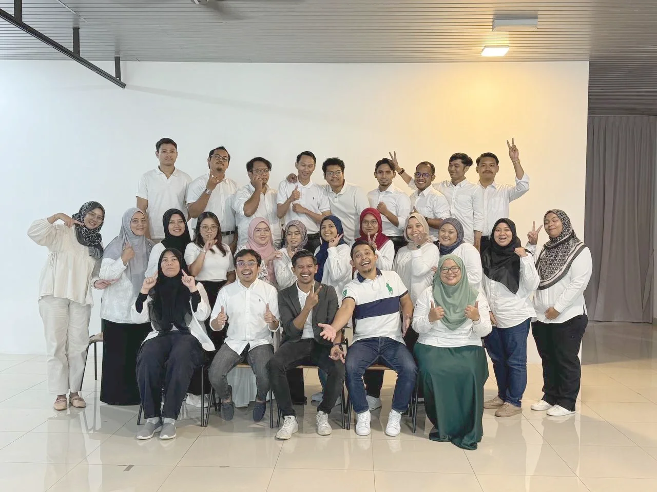 Group of diverse young adults posing for a photo indoors, some smiling and making gestures like peace signs, in a well-lit room with white walls and tiled floors.