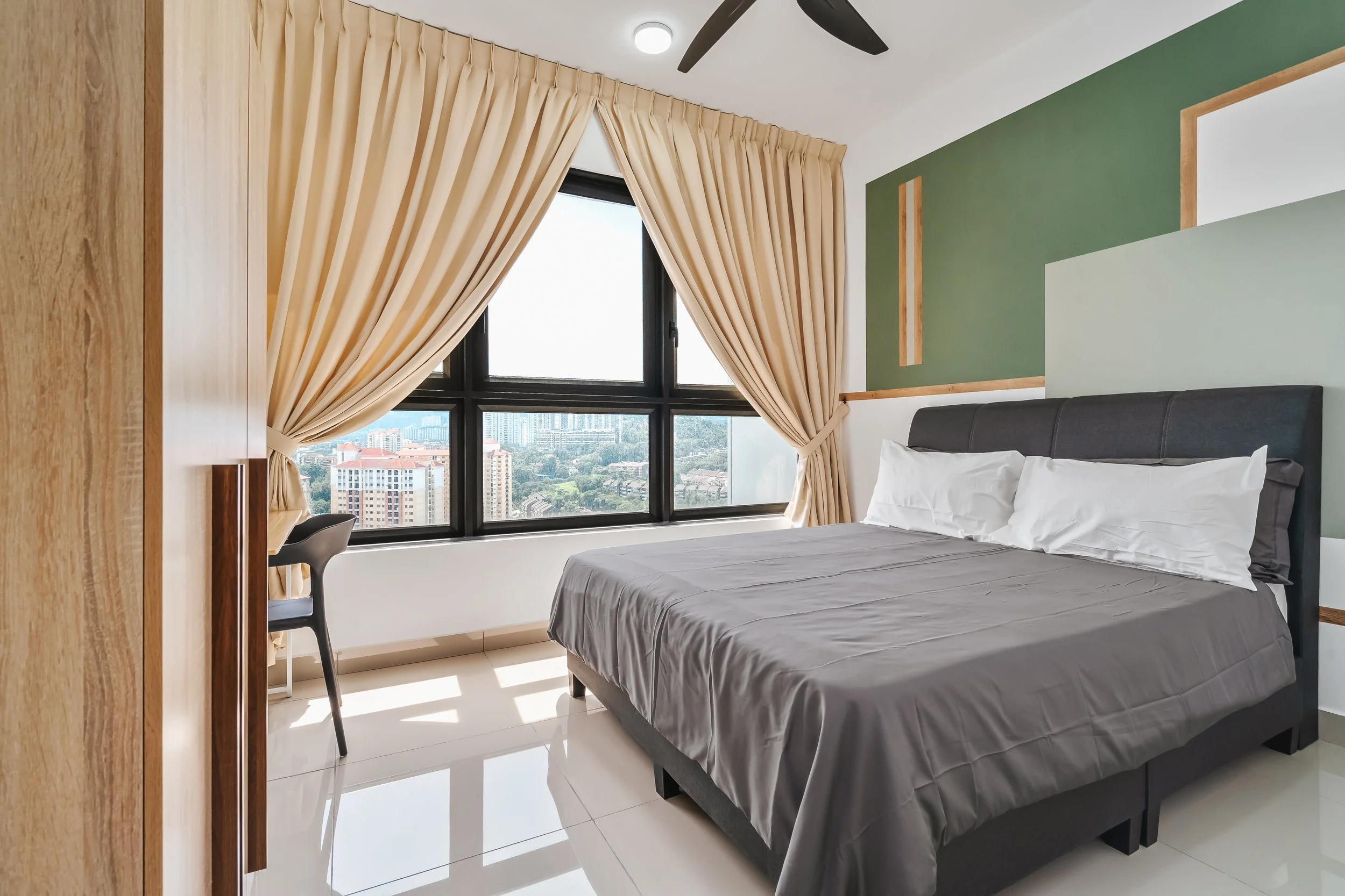 A bedroom with a large window and beige curtains, a black ceiling fan, a green accent wall with wood trim, a bed with gray bedding, a black headboard, and a small black chair.