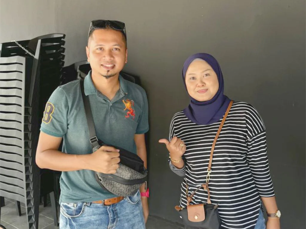 A man and a woman standing side by side indoors against a dark wall. The man is wearing a green polo shirt, sunglasses on his head, and a small bag across his chest. The woman is wearing a striped long-sleeve shirt, a navy blue hijab, and has a small