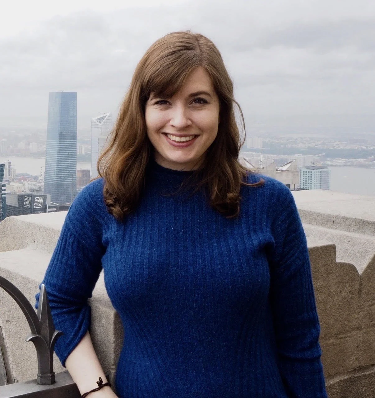 A woman with brown hair wearing a blue sweater, smiling on a rooftop with a city skyline and river in the background.