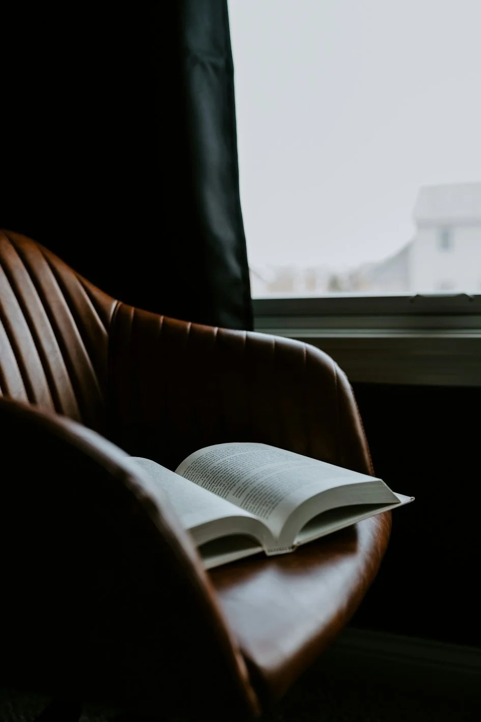 An open book resting on a brown leather armchair near a window with black curtains, with a cityscape visible outside.