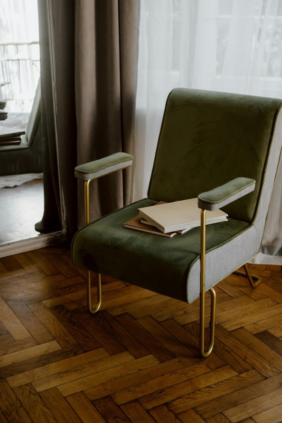 A green armchair with gold metal arms and legs, placed on a wooden parquet floor. There are several books or magazines on its seat. A sheer curtain and a glass door or window are behind the chair, with some outdoor space visible.
