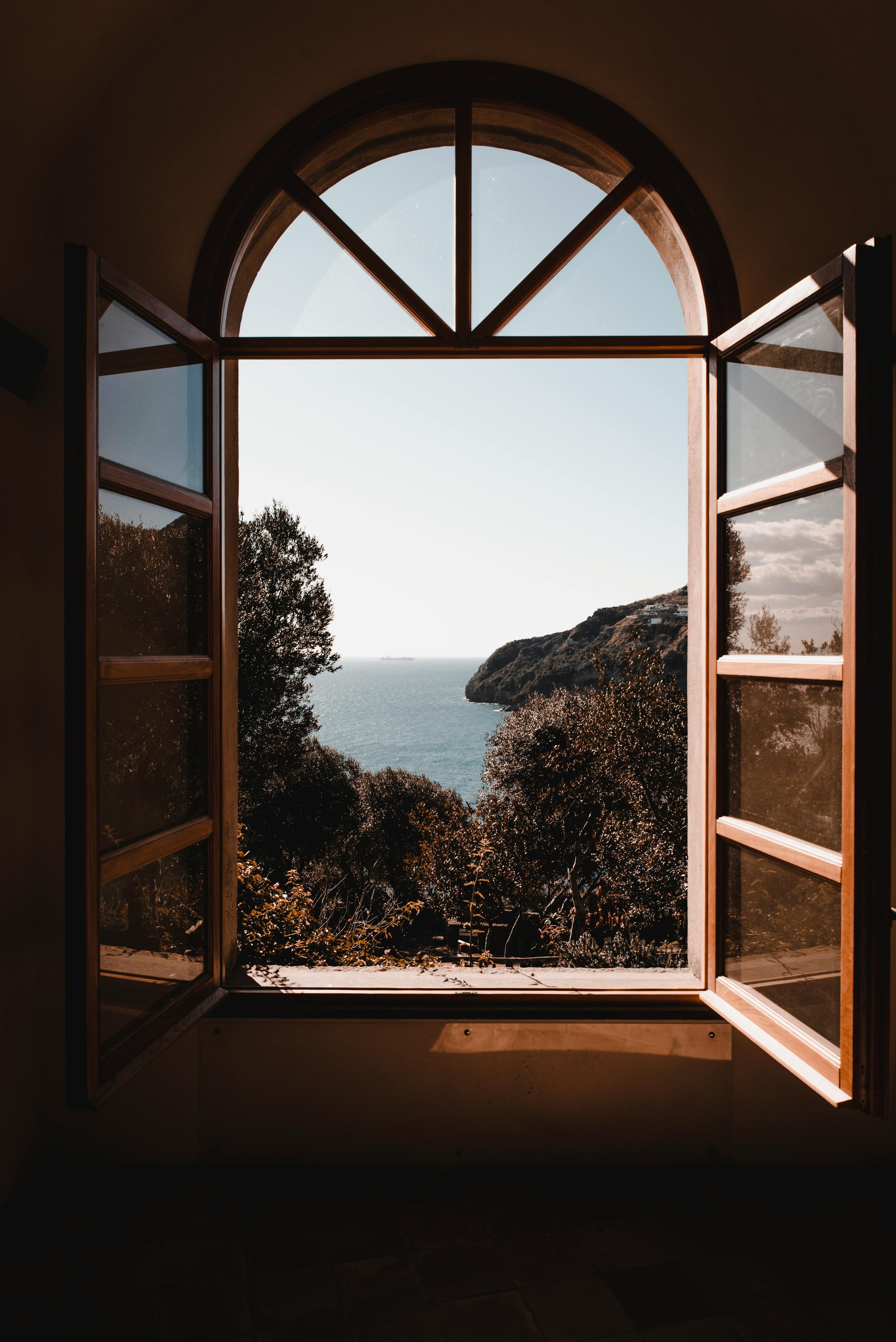 Open wooden window overlooking a coastal landscape with trees, ocean, and distant ship