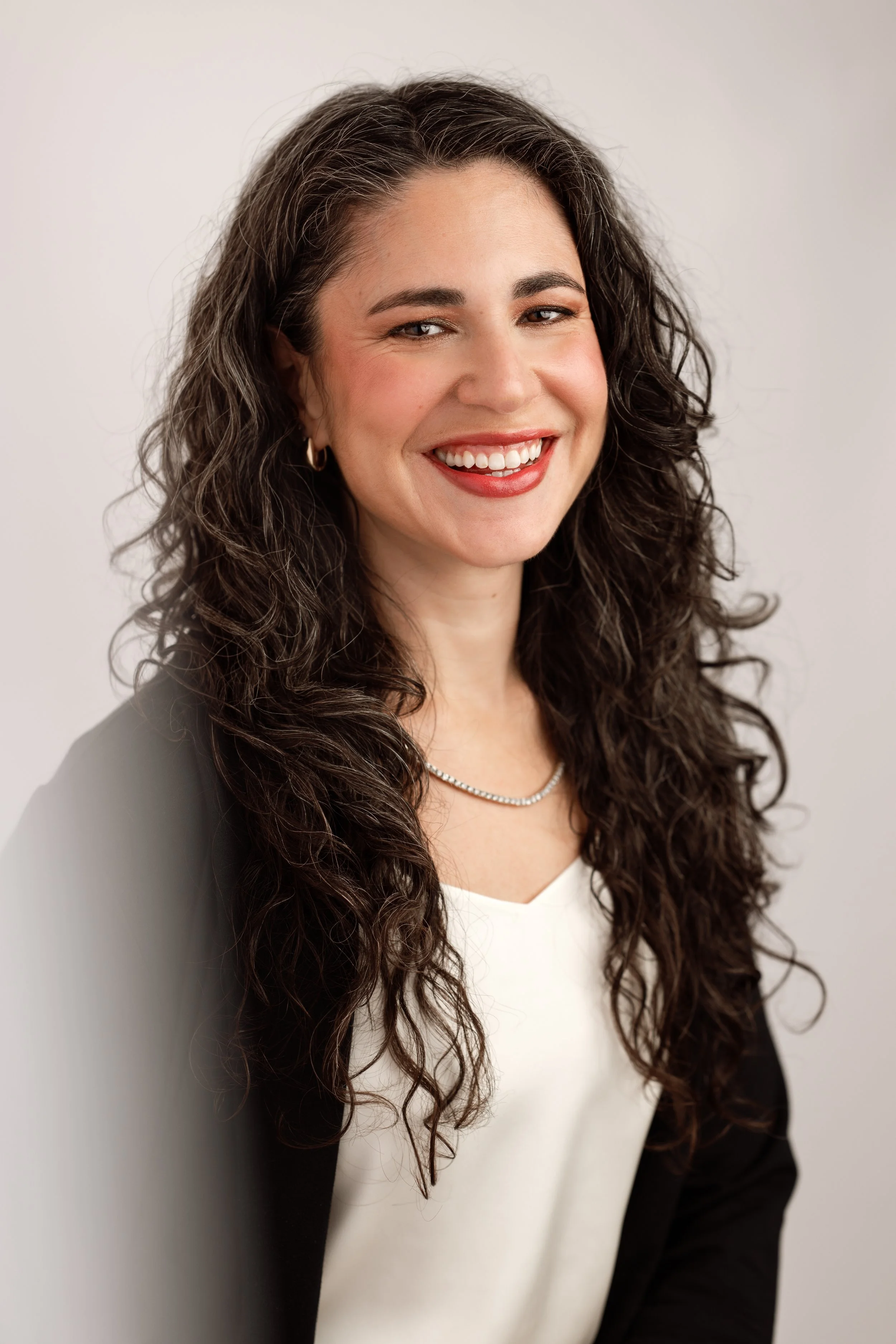 A smiling woman with long, curly dark hair wearing a black blazer, white blouse, and a pearl necklace, standing against a plain light background.