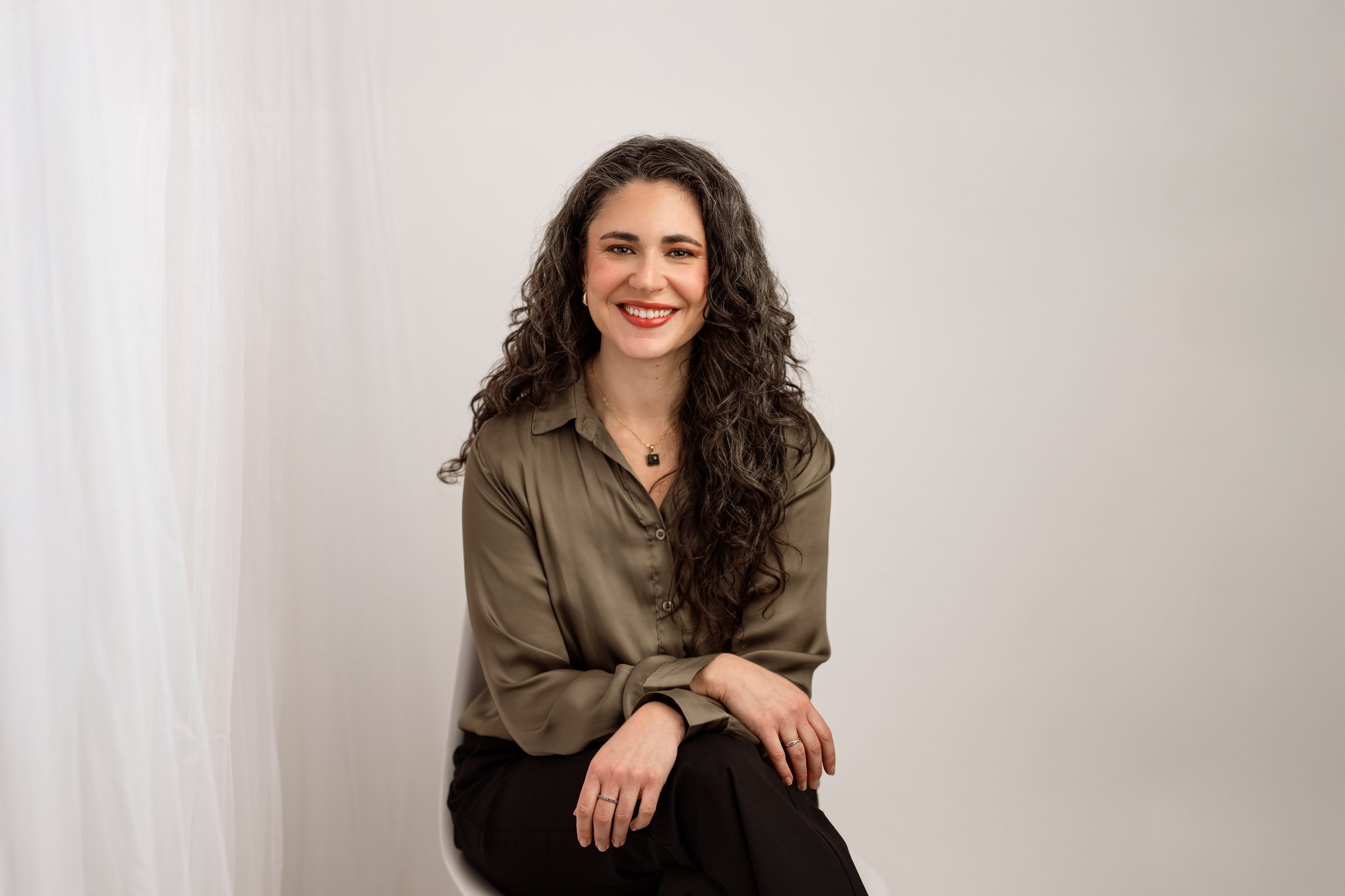 A woman with long curly dark hair and light skin sitting on a white chair in front of a plain off-white wall, smiling at the camera, wearing an olive green blouse and black pants.