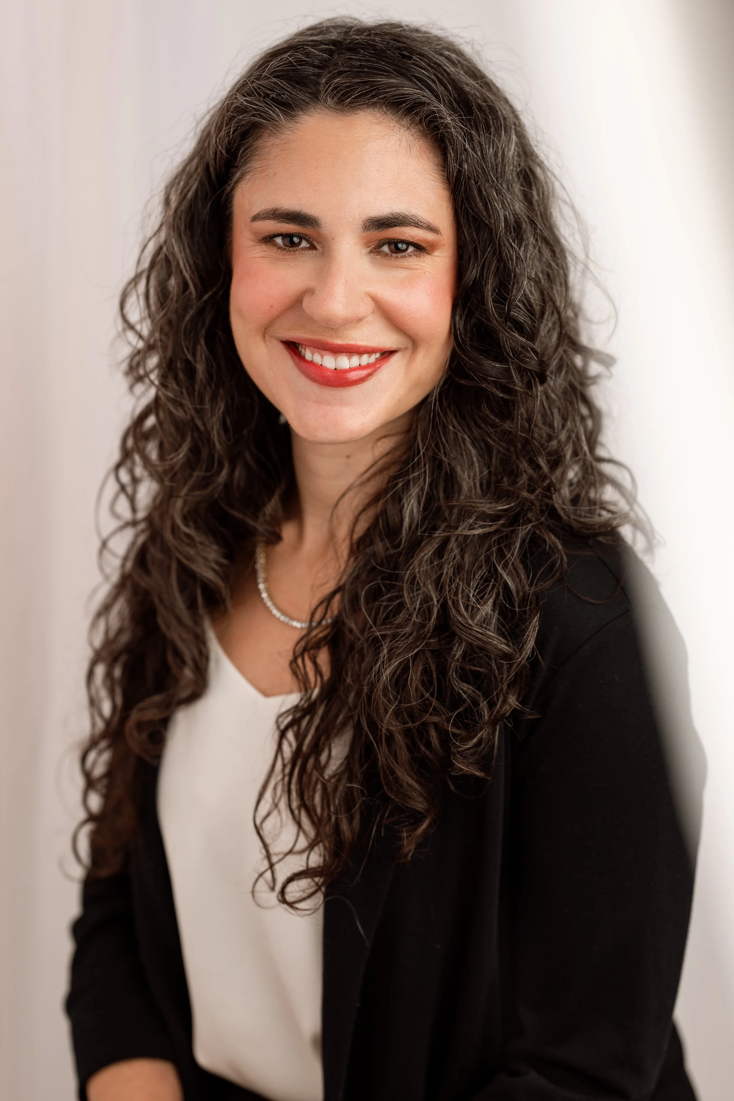 A woman with long, curly brown hair smiling, wearing a white blouse, black blazer, and a pearl necklace, standing against a neutral background.