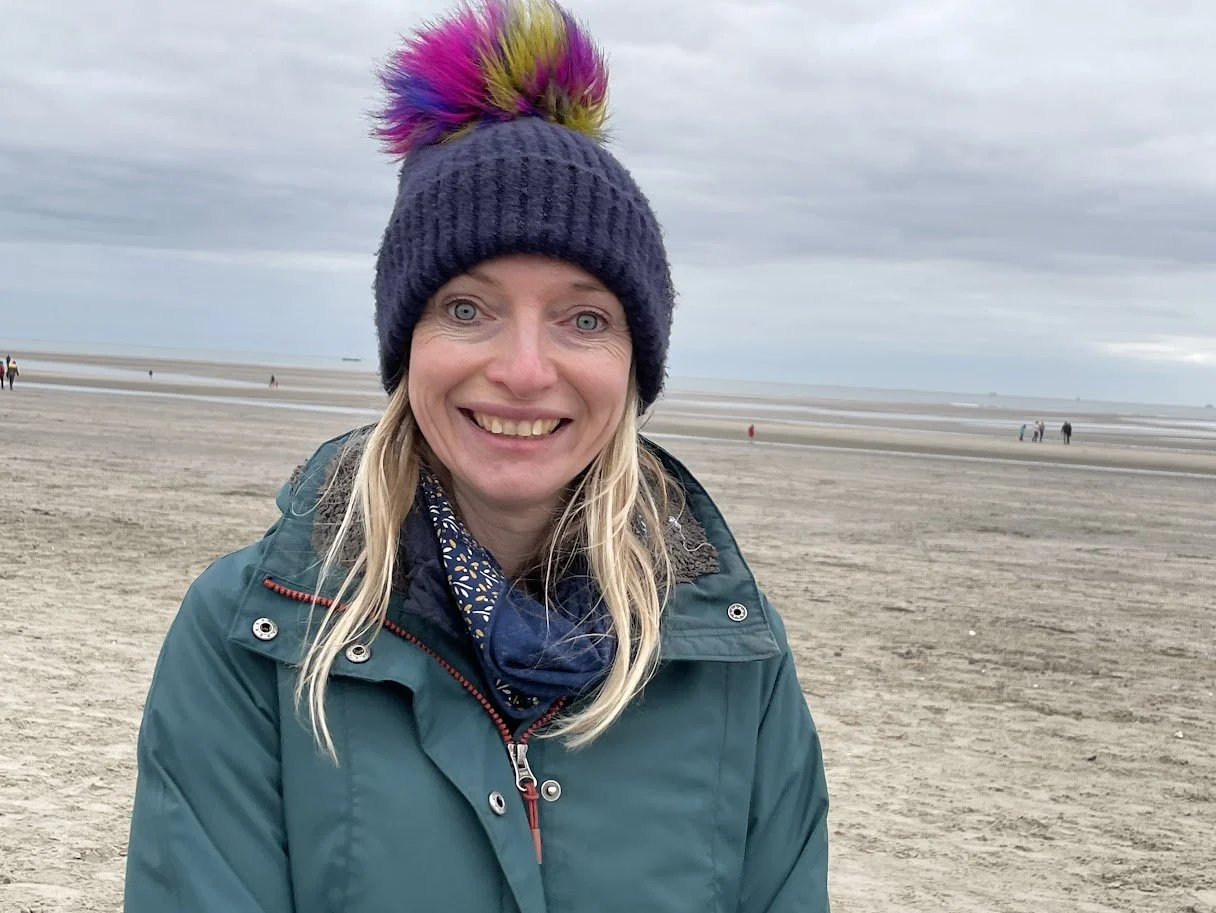 A woman smiling on a cloudy beach, wearing a knit hat with a colorful pom-pom, a teal jacket, and a patterned scarf.