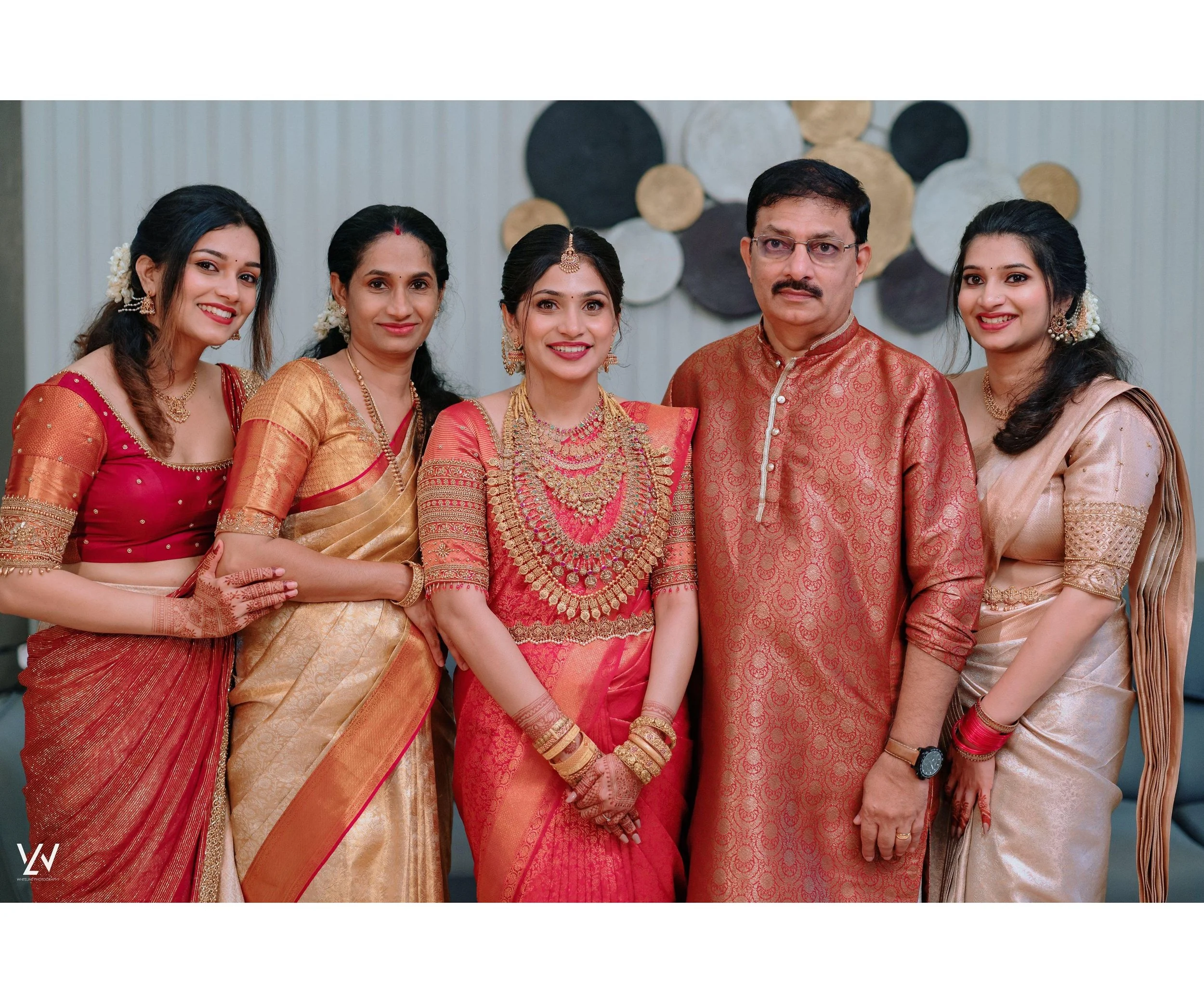 Family celebrating a traditional Indian wedding, with women in sarees and the man in a kurta, standing indoors with decorative wall art in the background.