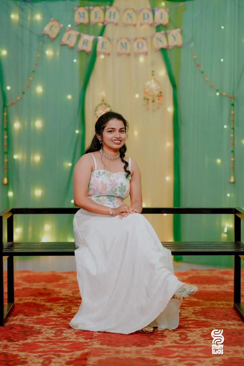 A young woman sitting on a black bench, dressed in a white dress with a floral top, smiling at the camera. The background features green and yellow curtains, fairy lights, and a festive banner that reads 'Mehndi & Lemon'.