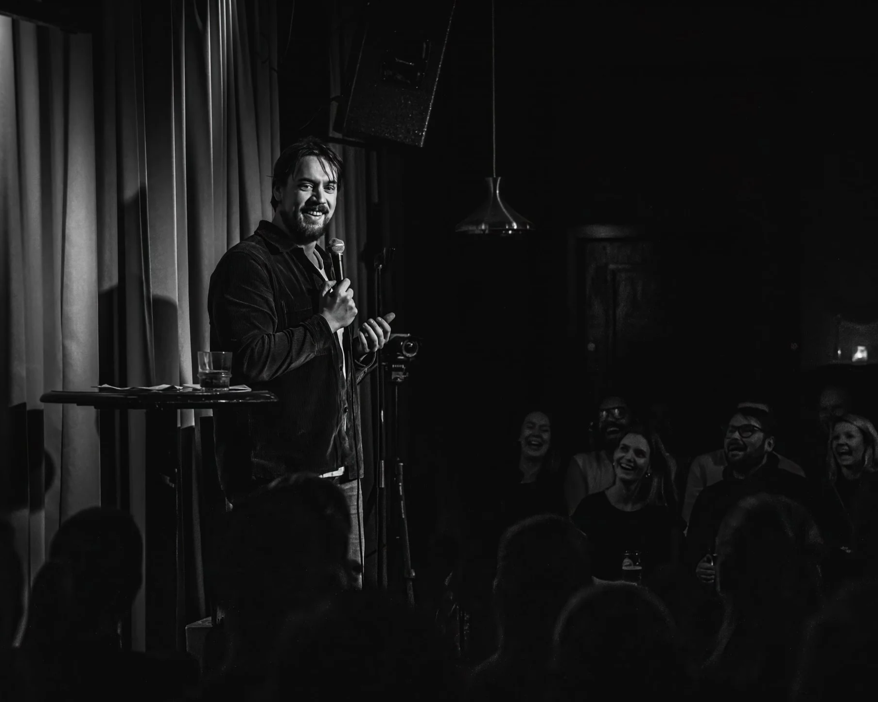 Komiker Espen Abrahamsen står på scenen og gjør standup for et publikum, smiler og holder en mikrofon i en svakt opplyst standupklubb.