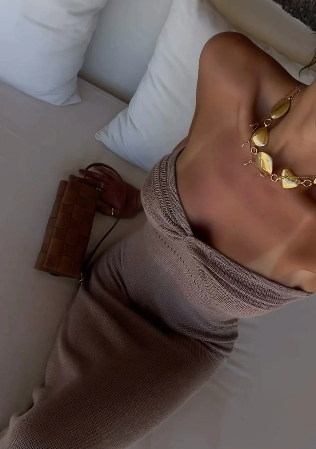 A woman lying on a bed wearing a beige sleeveless dress, a gold necklace, and with a brown quilted handbag beside her on the bed.