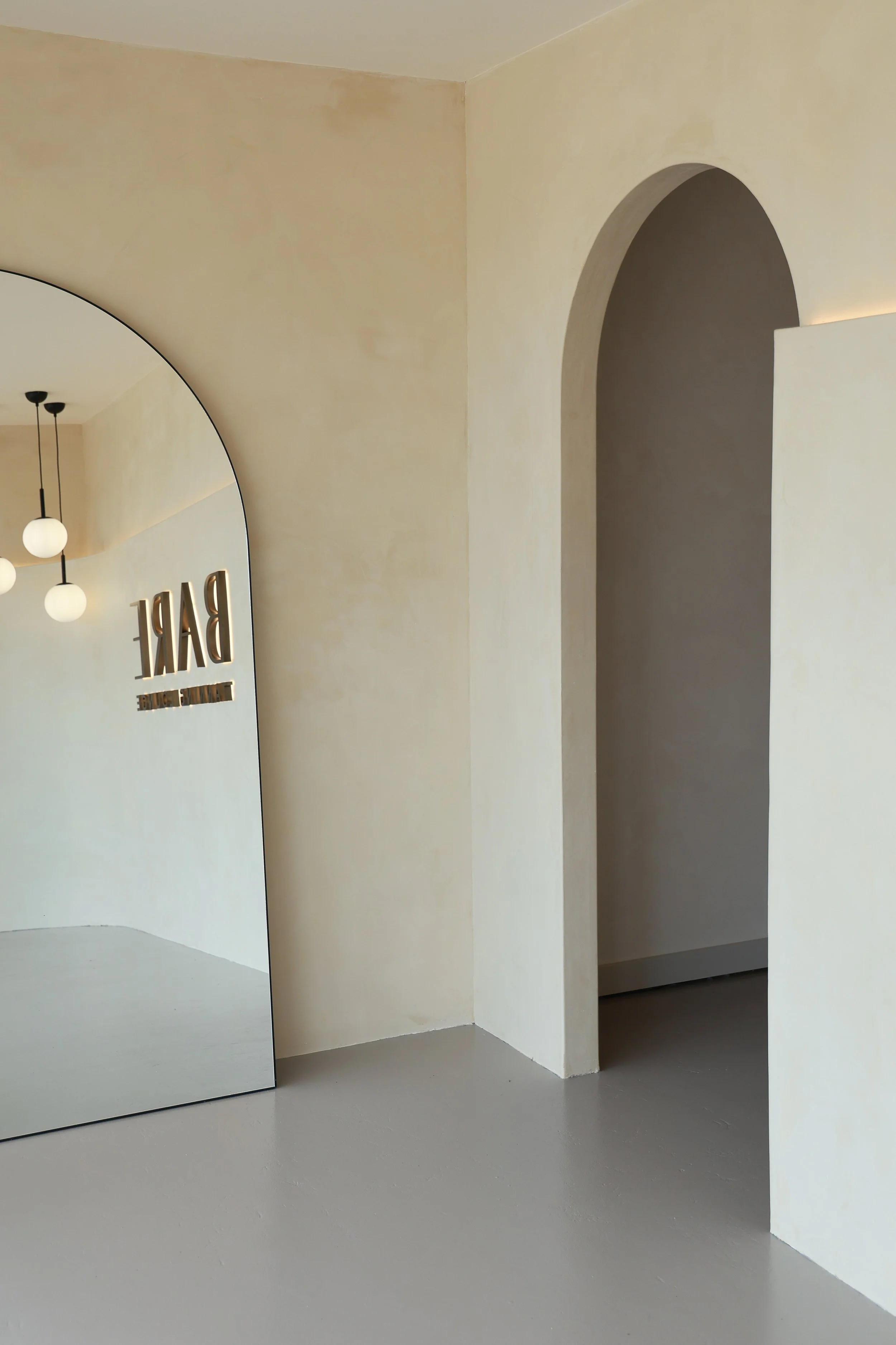 Minimalist interior space with a large, curved mirror on the left, a beige wall, pendant ceiling lights reflected in the mirror, and an arched doorway on the right.