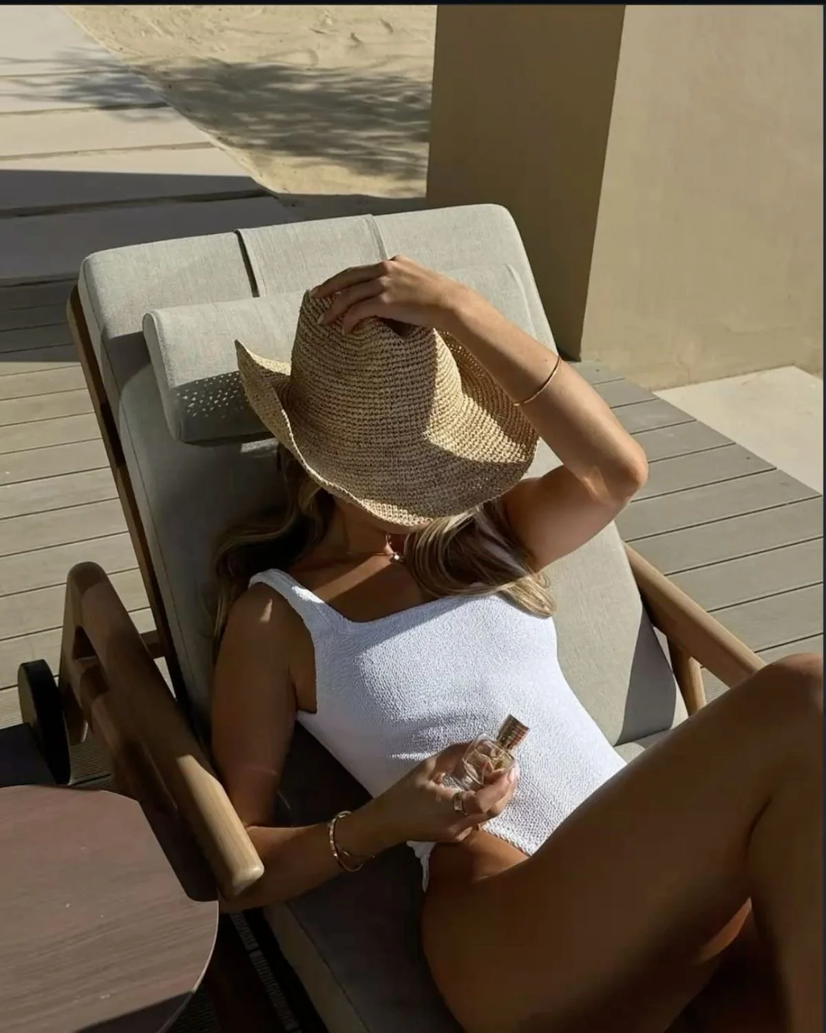 Woman lying on a lounge chair outdoors, wearing a white sleeveless top, holding a perfume bottle, and covering her face with a wide-brimmed straw hat.