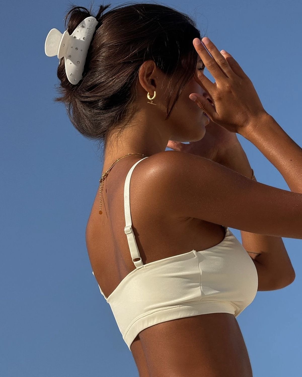 A woman with dark hair in a bun, wearing a white top and a large hair clip, is standing against a bright blue sky. She is holding her hand up to her face, with her eyes closed.