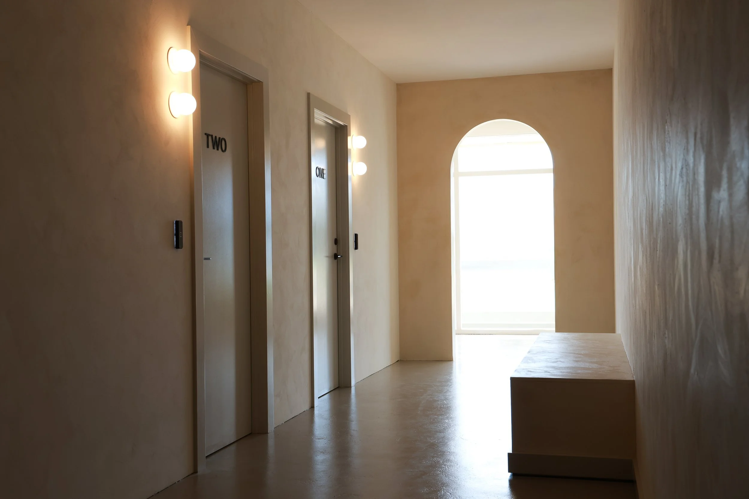 A hallway with two elevator doors labeled "ONE" and "TWO," beige textured walls, modern wall-mounted lights, and a bright arched window at the end of the corridor.