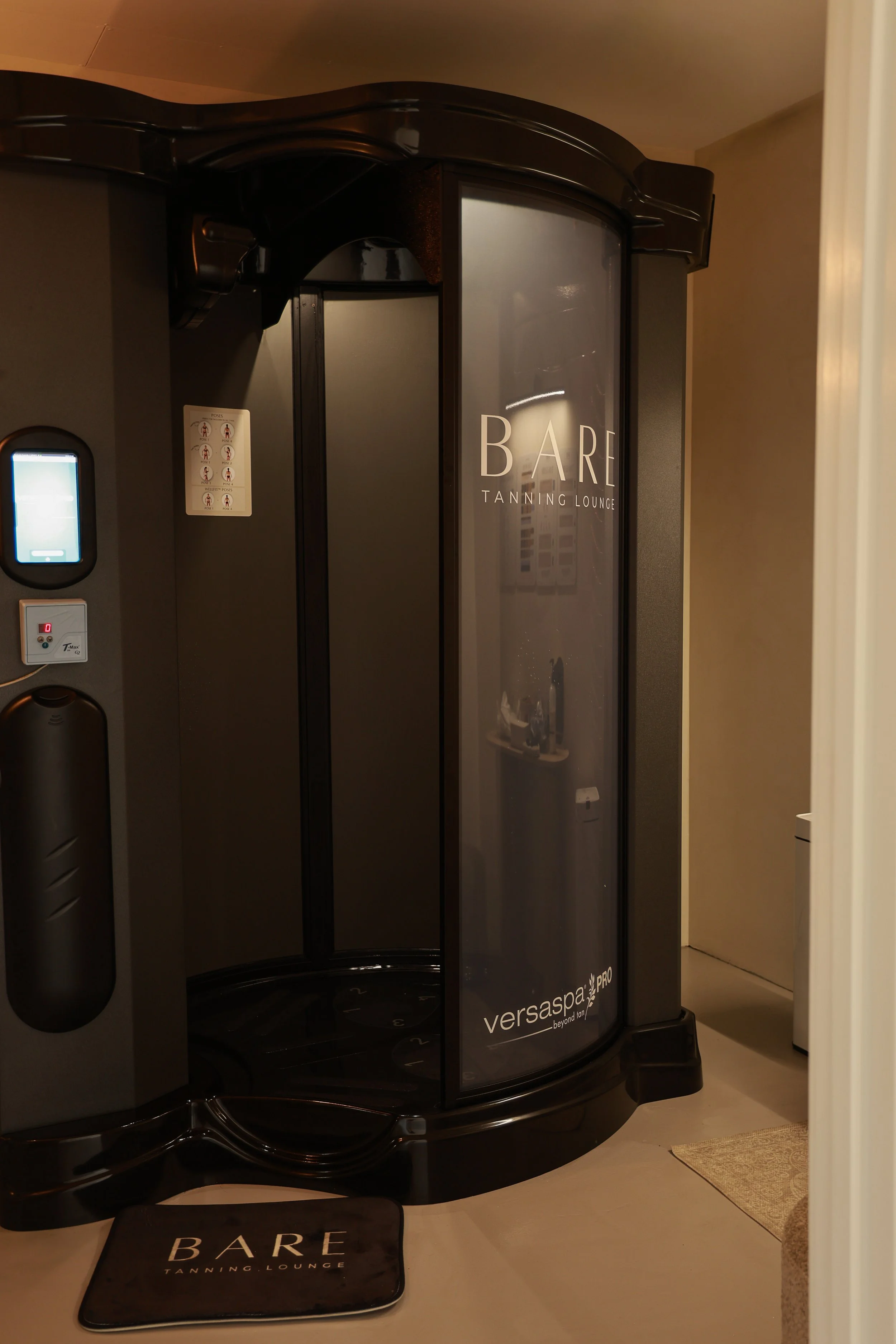 Indoor tanning capsule with glass door labeled 'BARE Tanning Lounge' and a mat outside reading 'BARE'.