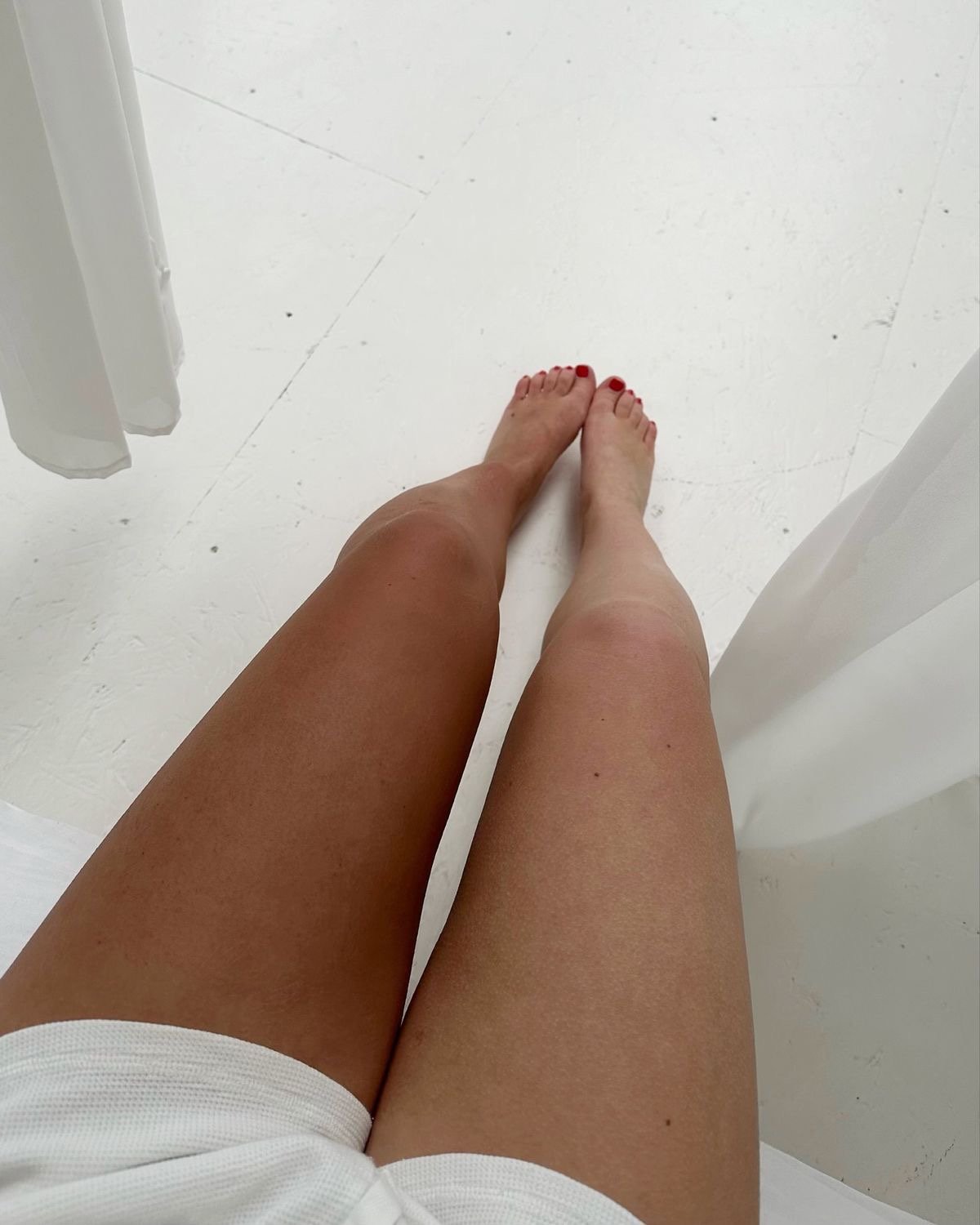A person's legs with tan skin and red toenail polish, standing on a white floor near white curtains.