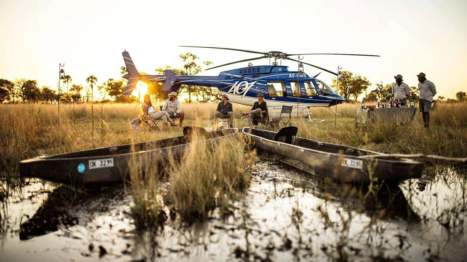 People sitting around a campfire near two boats with a helicopter in the background, set in a grassy field at sunset.