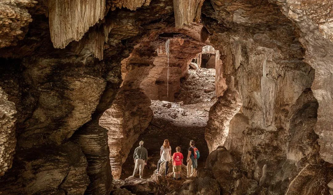 Group of five people exploring inside a large, natural cave with rugged rock formations and an opening leading outside.