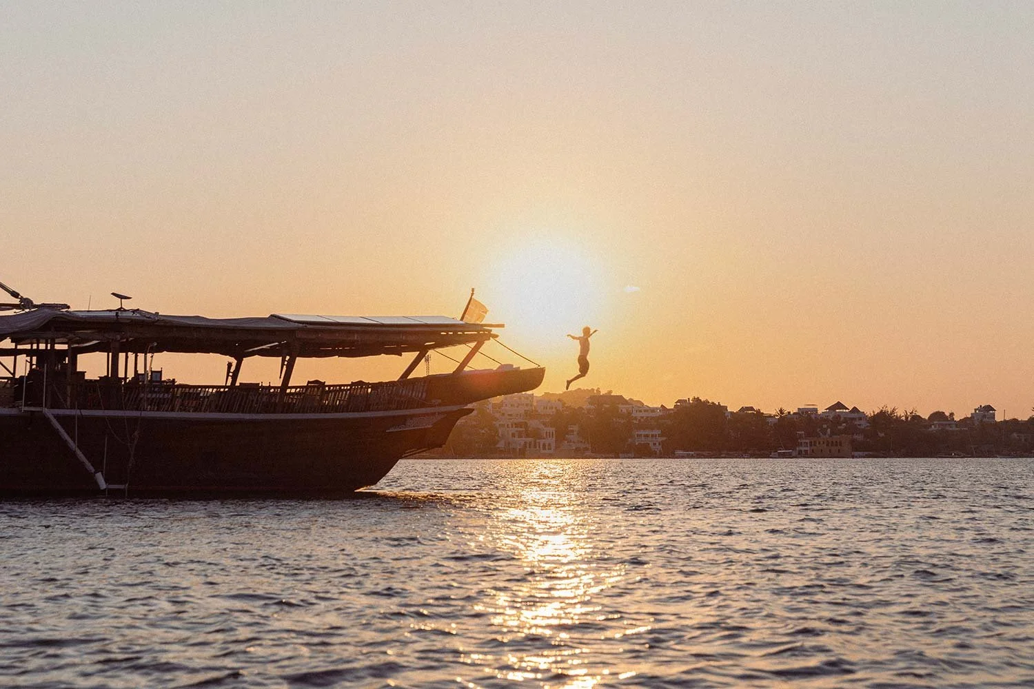 Person jumping off a boat into the water during sunset with buildings in the background.