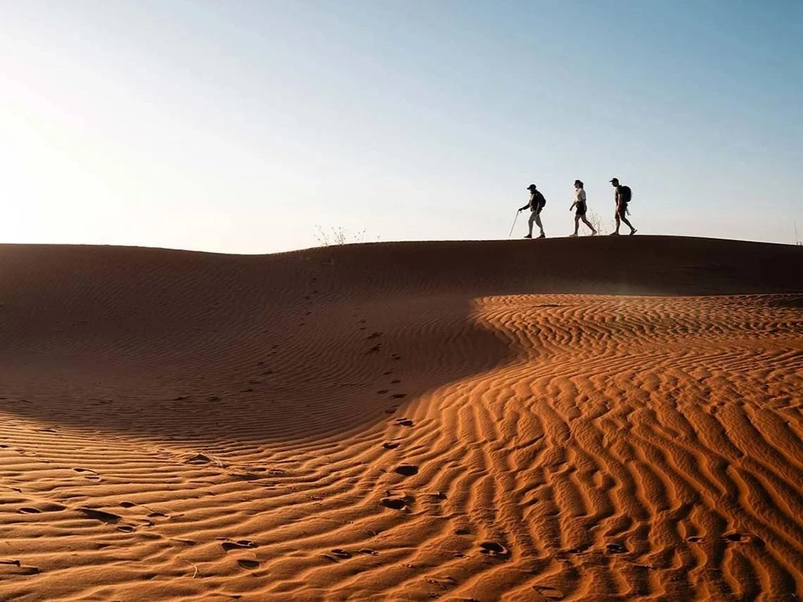 Three hikers with backpacks and walking sticks walking across a desert sand dune at sunset.