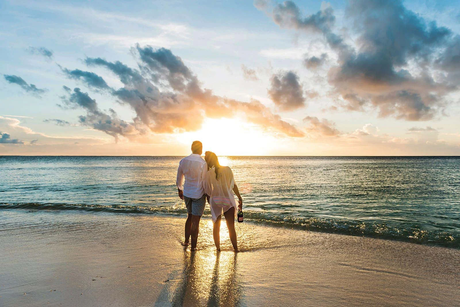 A couple walking along the beach at sunset, holding hands, with the ocean and clouds in the background.