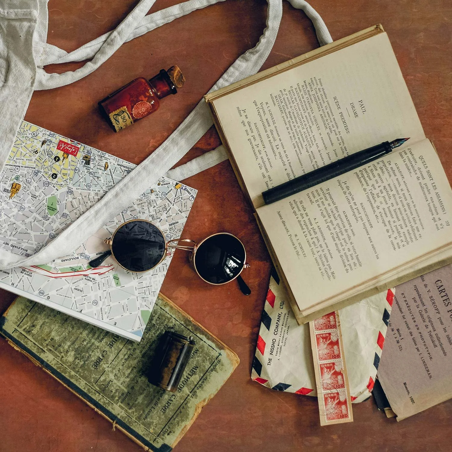 A cluttered wooden table with a map, vintage black glasses, an open book with a pen, a small bottle of medicine, a torn book, an envelope with postage stamps, and a white cloth strap.