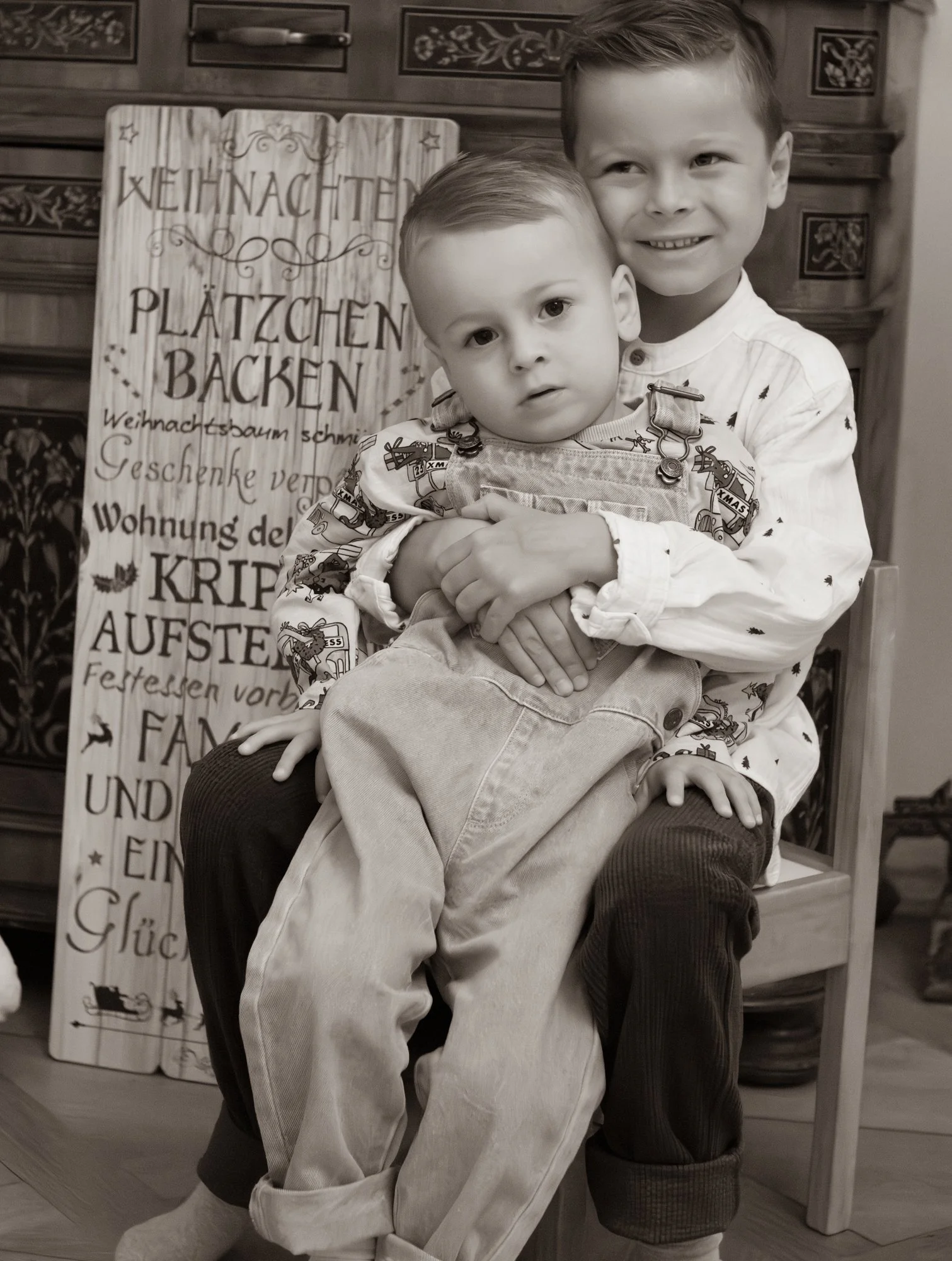 Zwei Jungen, ein älterer und ein jüngerer, sitzen auf einem Stuhl, das ältere Kind umarmt das jüngere, im Hintergrund ein Weihnachtsdekorationsschild mit dem Wort 'Weihnachten' in Deutsch.