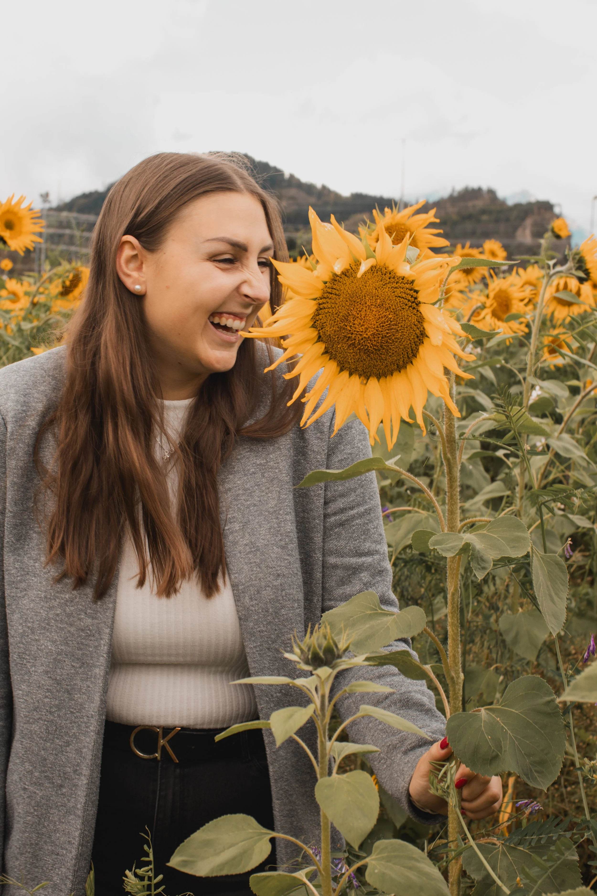 Eine lachende Frau mit braunen Haaren in einem Sonnenblumenfeld, die eine Sonnenblume hält.